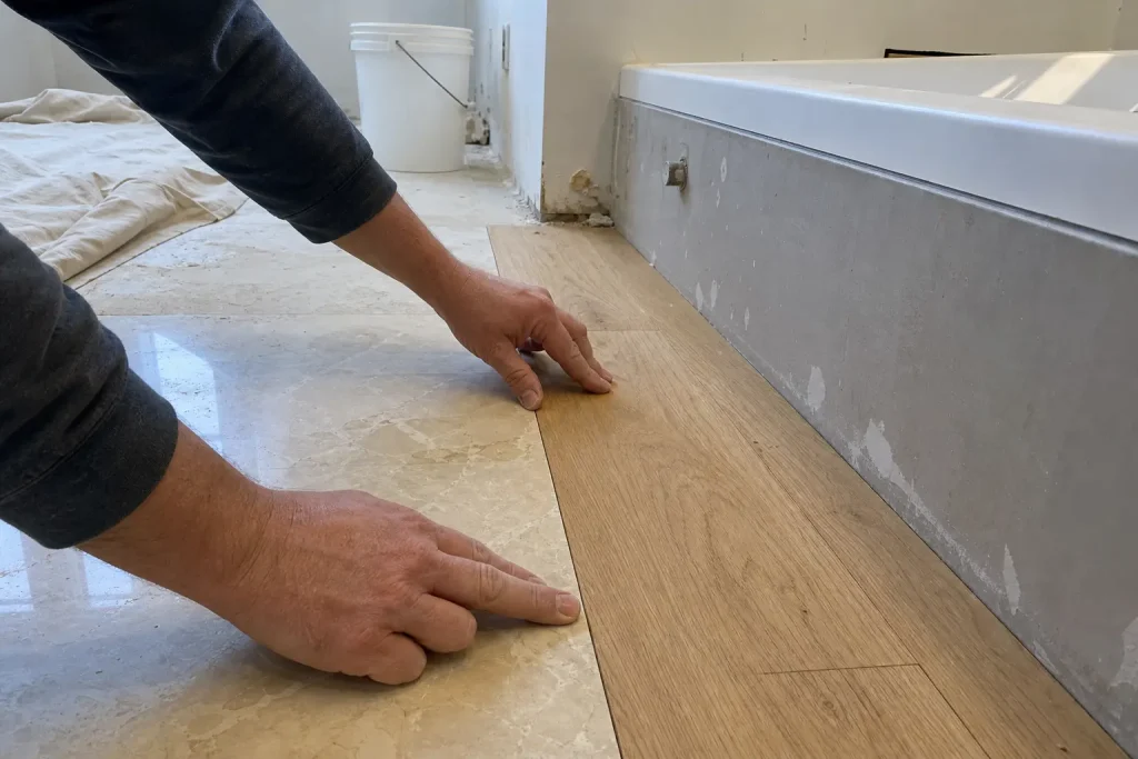 Händer som testar naturstensplatta och trägolv sida vid sida under badrumsrenovation i lantlig stil